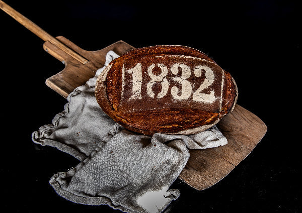 Ein rundes, längliches, braun gebackenes Brot liegt auf einem Holzbrett und einem grauen Tuch. Auf der Oberfläche ist die Zahl "1832" in weißer Schrift zu sehen