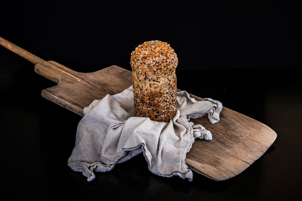 Ganzes, längliches Proteinbrot mit goldbrauner, dicht mit Saaten bestreuter Kruste, aufrecht auf einem rustikalen Holzbrett und einem hellen Leinentuch arrangiert.