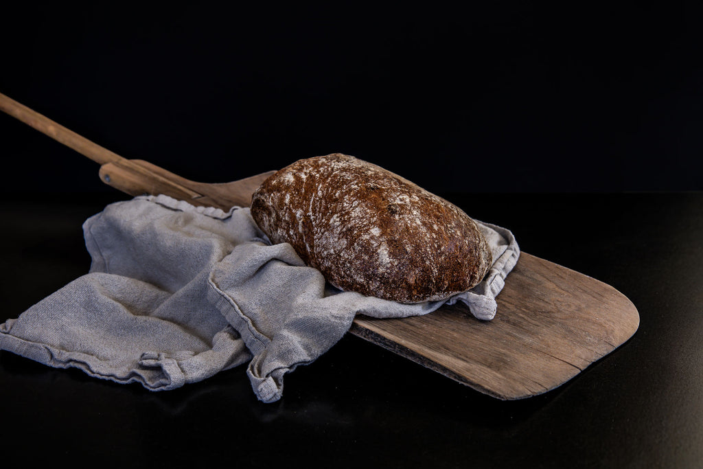 Rustikales längliches Bauernbrot mit kräftig gebräunter, bemehlter Kruste liegt auf einem Holzbrett und ist teilweise auf einem hellen Leinentuch arrangiert