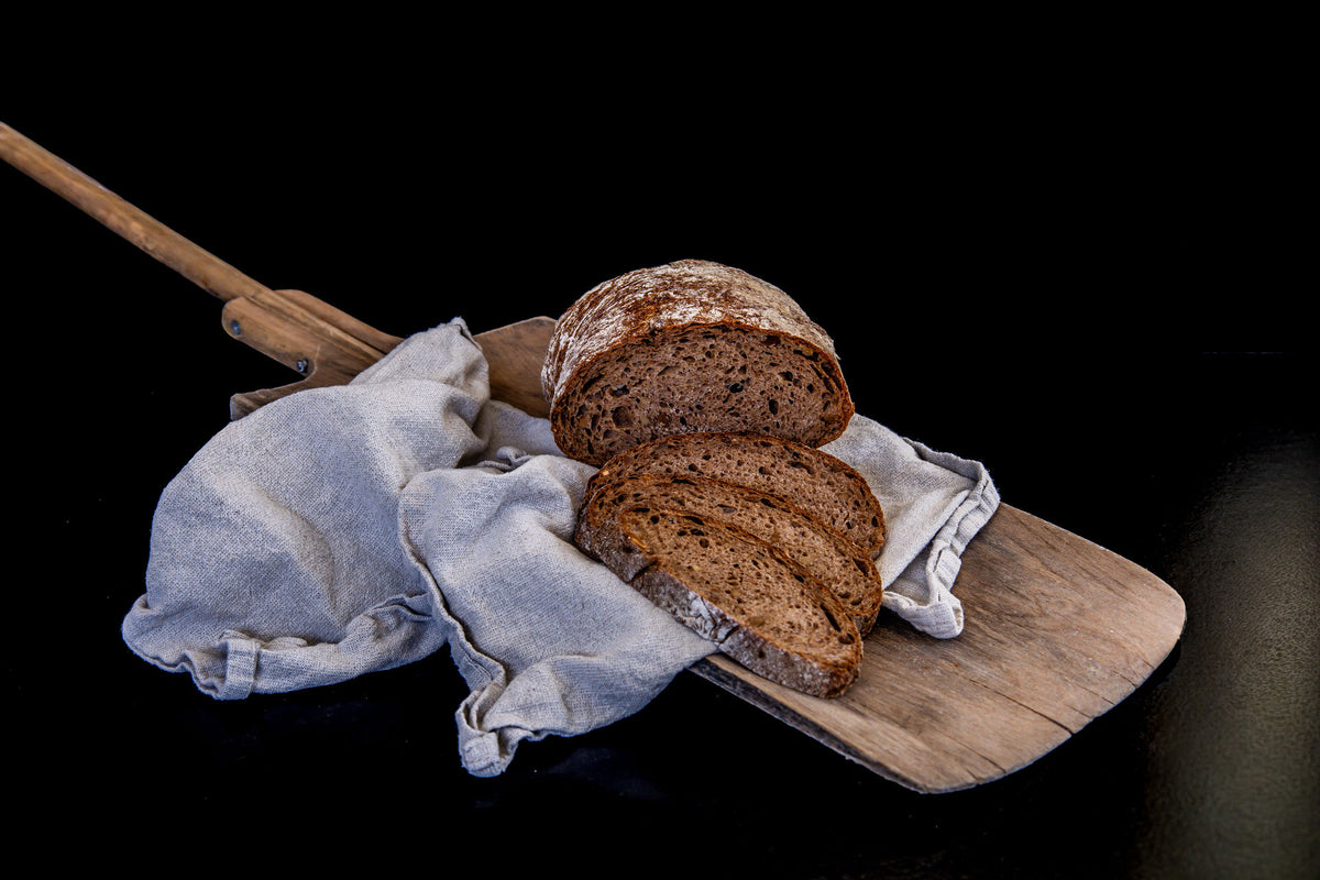 Aufgeschnittenes, rustikales Vollkornbrot mit kräftig gebräunter, bemehlter Kruste. Drei Brotscheiben liegen vor dem angeschnittenen Brot auf einem Holzbrett mit hellem Leinentuch