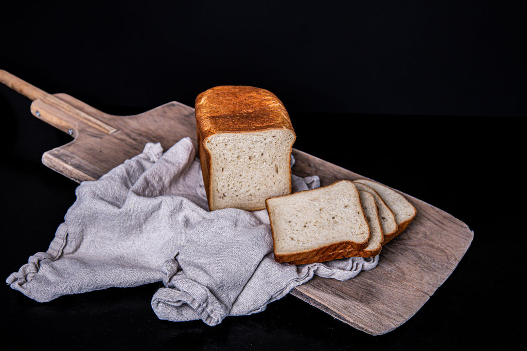 Kastenförmiges Weizenbrot mit goldbrauner Kruste, aufgeschnitten und mit mehreren Brotscheiben auf einem Holzbrett und hellen Leinentuch angerichtet