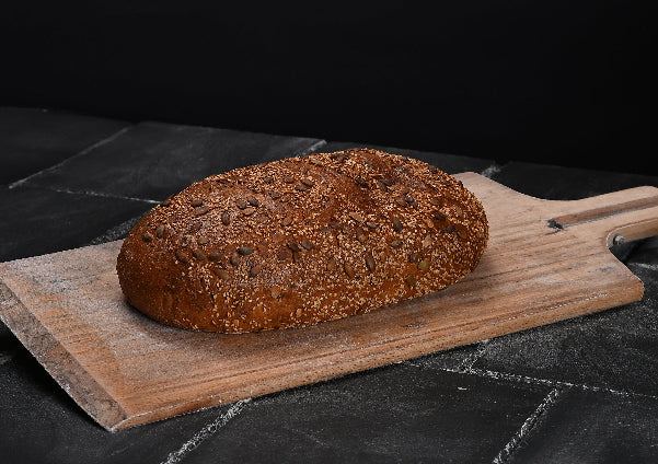 Flaches, ovales Mehrkornbrot mit goldbrauner, dicht mit verschiedenen Saaten bestreuter Kruste liegt auf einem hellen Holzbrett
