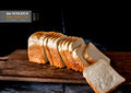 Geschnittener Toast liegt auf einem rustikalen Holzbrett. Mehrere Scheiben sind aufgefächert, eine Scheibe liegt vorne auf dem Brett. Im Hintergrund ist eine dunkle Fläche mit Mehlspuren zu sehen. Oben links ist das Logo der Bäckerei Schleich eingefügt.