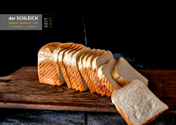 Geschnittener Toast liegt auf einem rustikalen Holzbrett. Mehrere Scheiben sind aufgefächert, eine Scheibe liegt vorne auf dem Brett. Im Hintergrund ist eine dunkle Fläche mit Mehlspuren zu sehen. Oben links ist das Logo der Bäckerei Schleich eingefügt.