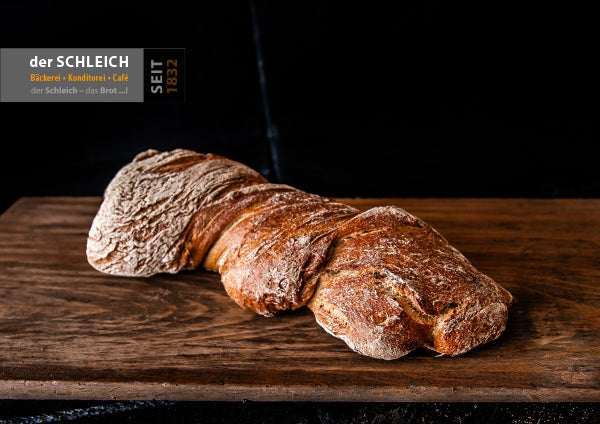 Ein rustikales, längliches Bauernbrot mit kräftiger, bemehlter Kruste liegt auf einem Holzbrett. Das Brot ist in rustikal eingerollt, damit es ausschaut wie eine Wurzel