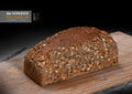 Ein längliches, goldbraunes Körnerbrot mit einer dicht mit Haferflocken und verschiedenen Körnern bedeckten Kruste liegt auf einem Holzbrett.