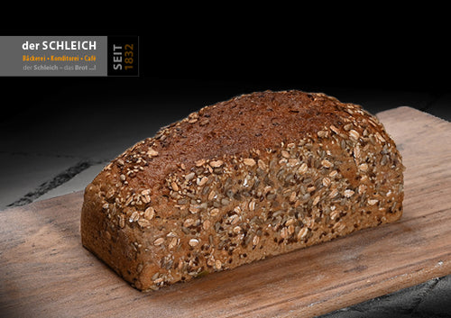 Ein längliches, goldbraunes Körnerbrot mit einer dicht mit Haferflocken und verschiedenen Körnern bedeckten Kruste liegt auf einem Holzbrett.