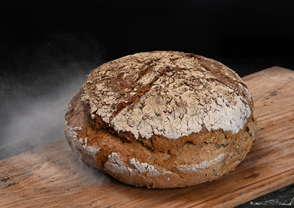 Ein rundes, rustikales Bauernbrot mit kräftiger, rissiger und bemehlter Kruste liegt auf einem Holzbrett. Das Brot ist goldbraun gebacken und hat eine unregelmäßige, aufgerissene Oberfläche auf der grobe Gewürze zu sehen sind.