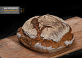 Ein rundes, rustikales Bauernbrot mit kräftiger, rissiger und bemehlter Kruste liegt auf einem Holzbrett. Das Brot ist goldbraun gebacken und hat eine unregelmäßige, aufgerissene Oberfläche.