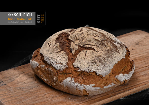 Ein rundes, rustikales Bauernbrot mit kräftiger, rissiger und bemehlter Kruste liegt auf einem Holzbrett. Das Brot ist goldbraun gebacken und hat eine unregelmäßige, aufgerissene Oberfläche.