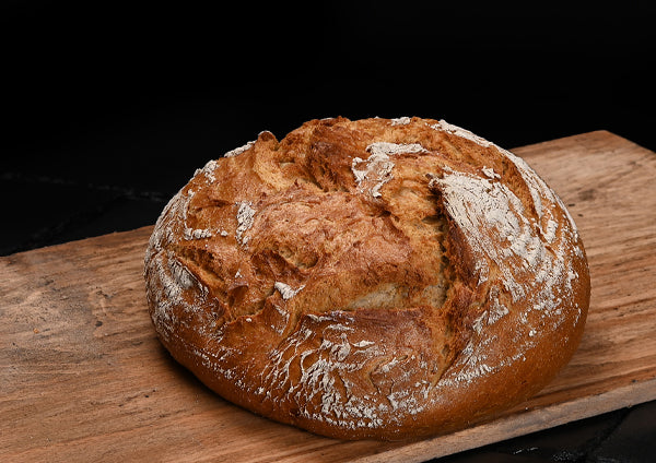Ein rundes, rustikales Bauernbrot mit goldbrauner, aufgerissener und teilweise bemehlter Kruste liegt auf einem Holzbrett