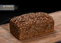 Ein längliches, goldbraunes Körnerbrot mit einer dicht mit Sonnenblumenkernen bedeckten Kruste liegt auf einem Holzbrett.