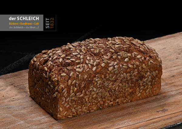 Ein längliches, goldbraunes Körnerbrot mit einer dicht mit Sonnenblumenkernen bedeckten Kruste liegt auf einem Holzbrett.